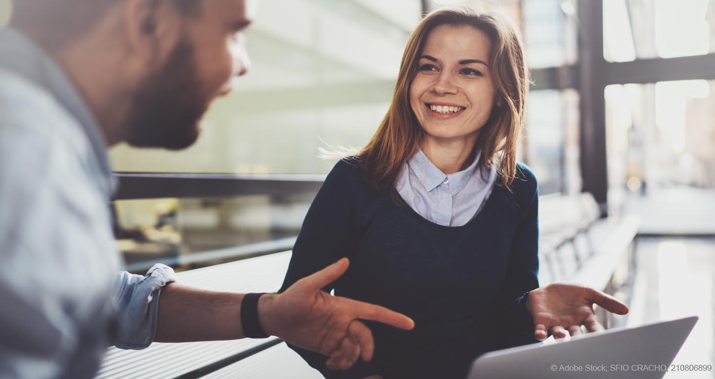 Eine junge Frau und ein junger Mann diskutieren im Arbeitskontext miteinander. Die Frau hat einen PC bei sich und lächelt.