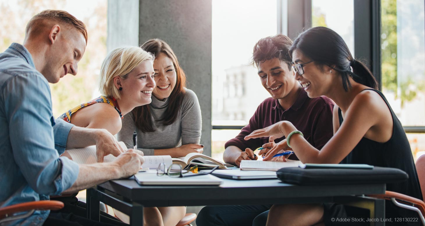 Eine Gruppe fünf junger Erwachsener sitzt am Tisch lernen und tauschen sich aus. Auf dem Tisch liegen Bücher und Notebooks
